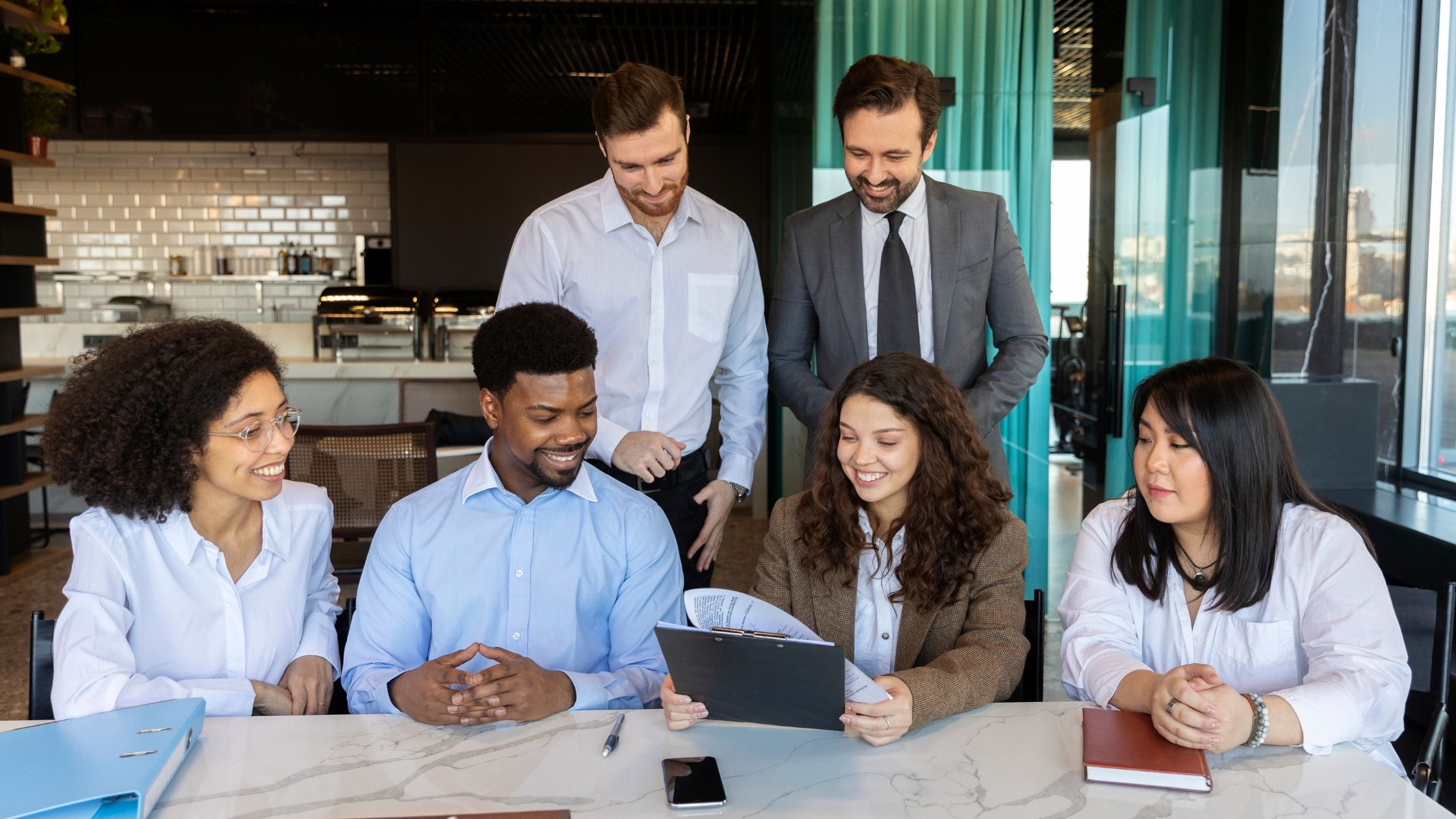 A diverse group of six professionals gathers around a conference table in a bright, modern space, smiling and reviewing documents together with energy and engagement.