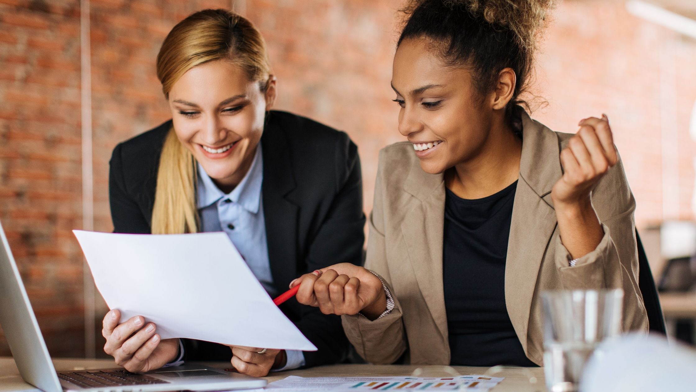 Two professionals review a printed report together at a desk, both smiling with visible energy and engagement — one pointing to the document with a pen, the other leaning in with an enthusiastic reaction, suggesting a positive outcome from the discussion.