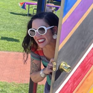 Nina Schiarizzi-Tobin wearing white sunglasses "coming out" of a rainbow door