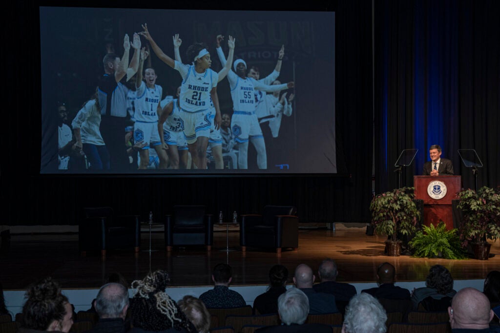 The stage at the State of the University event, with President Marc Parlange at the podium. A photo of the women's basketball team cheering is projected on a large screen in the background.