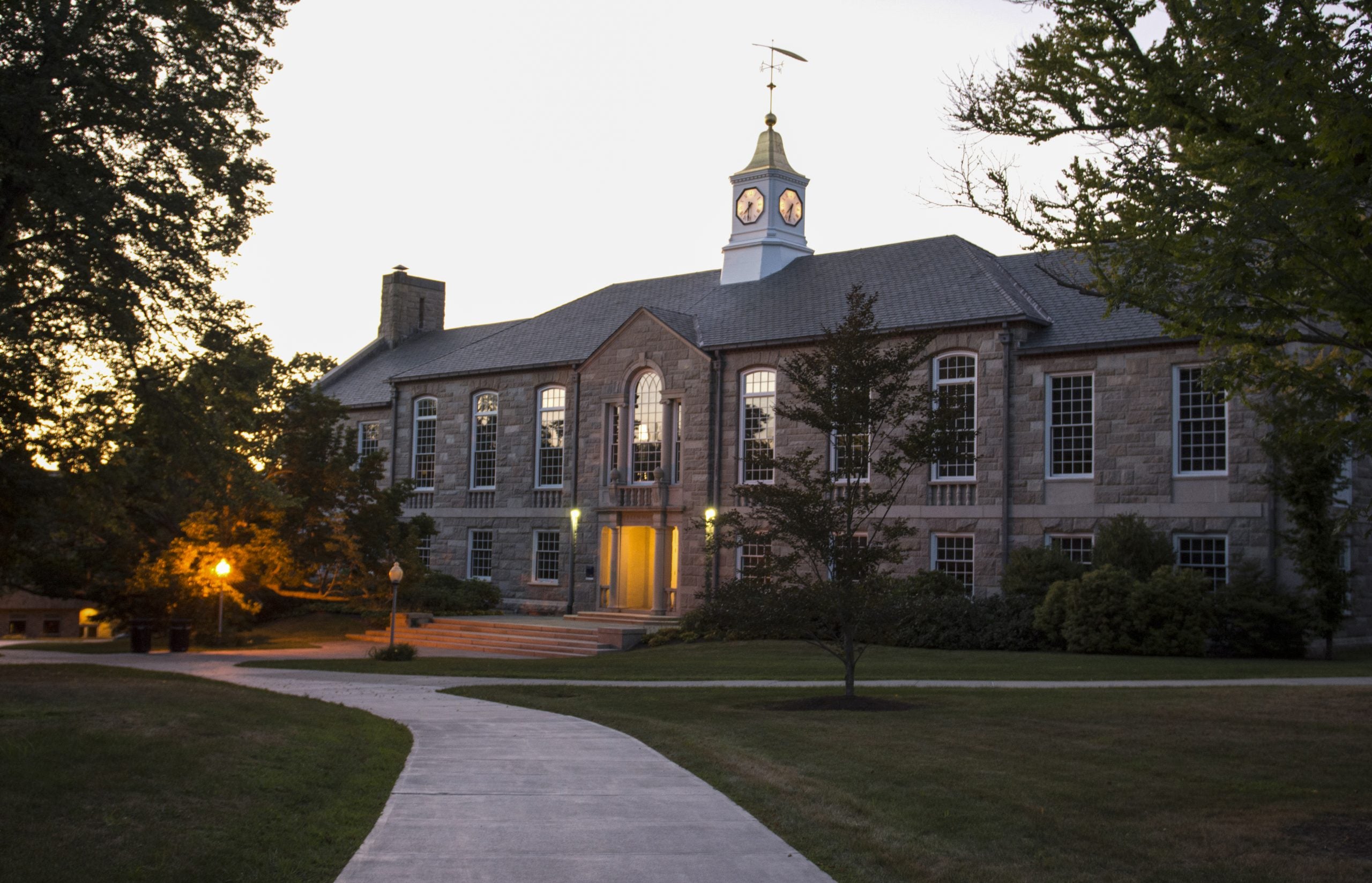 Green Hall, a stone building at the University of Rhode Island, pictured at night with the sunset above