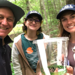 Paul Roselli, president of the Burrillville Land Trust; URI research assistant Saffron Zaniewski, and Alana Russell, manager of the URI Biocontrol Lab, at the Edward Vock Conservation Area in Pascoag holding a container of 209 parasitoid wasps in hopes to protect local ash trees from extinction.