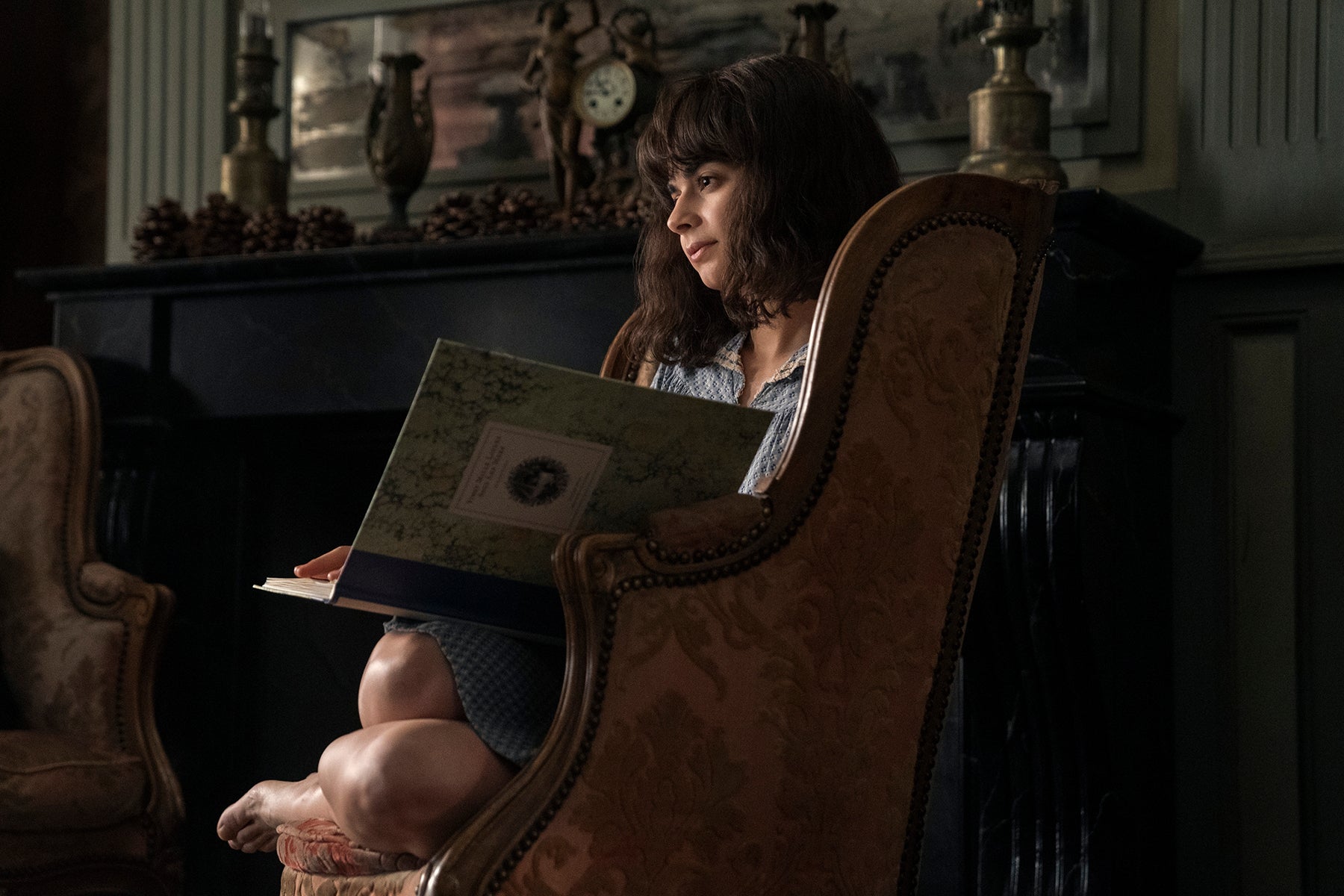 Aria Mia Loberti sitting in a chair looking to the left, holding a book open in her lap