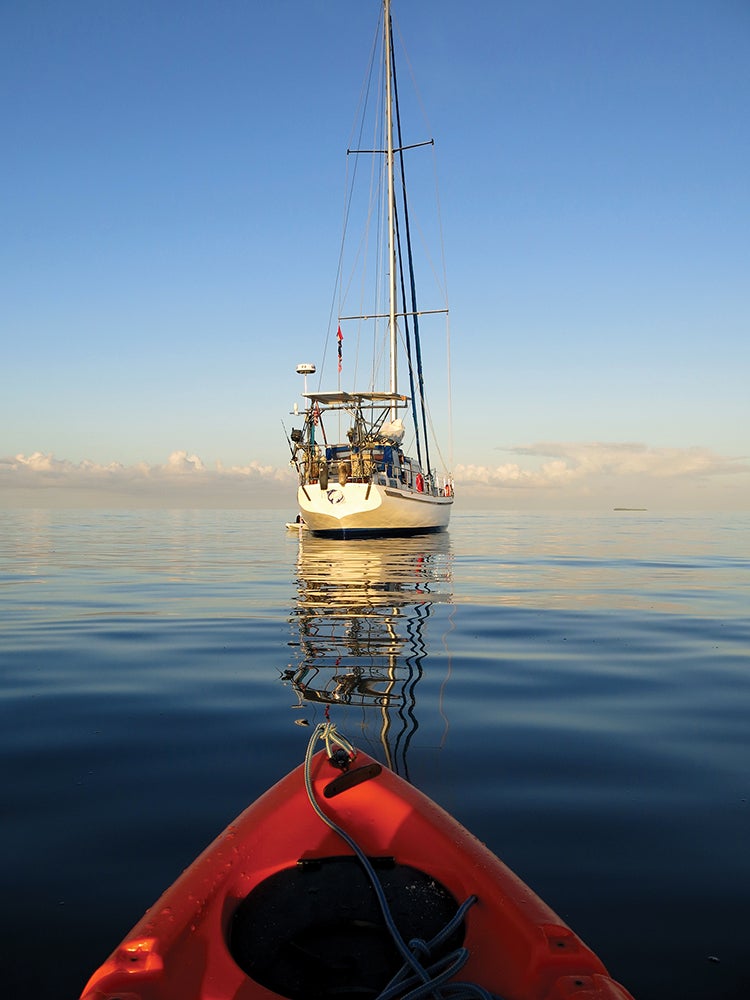 The prow of a kayak follows a yacht on the water with a clear blue sky except for a row of clouds on the horizon.