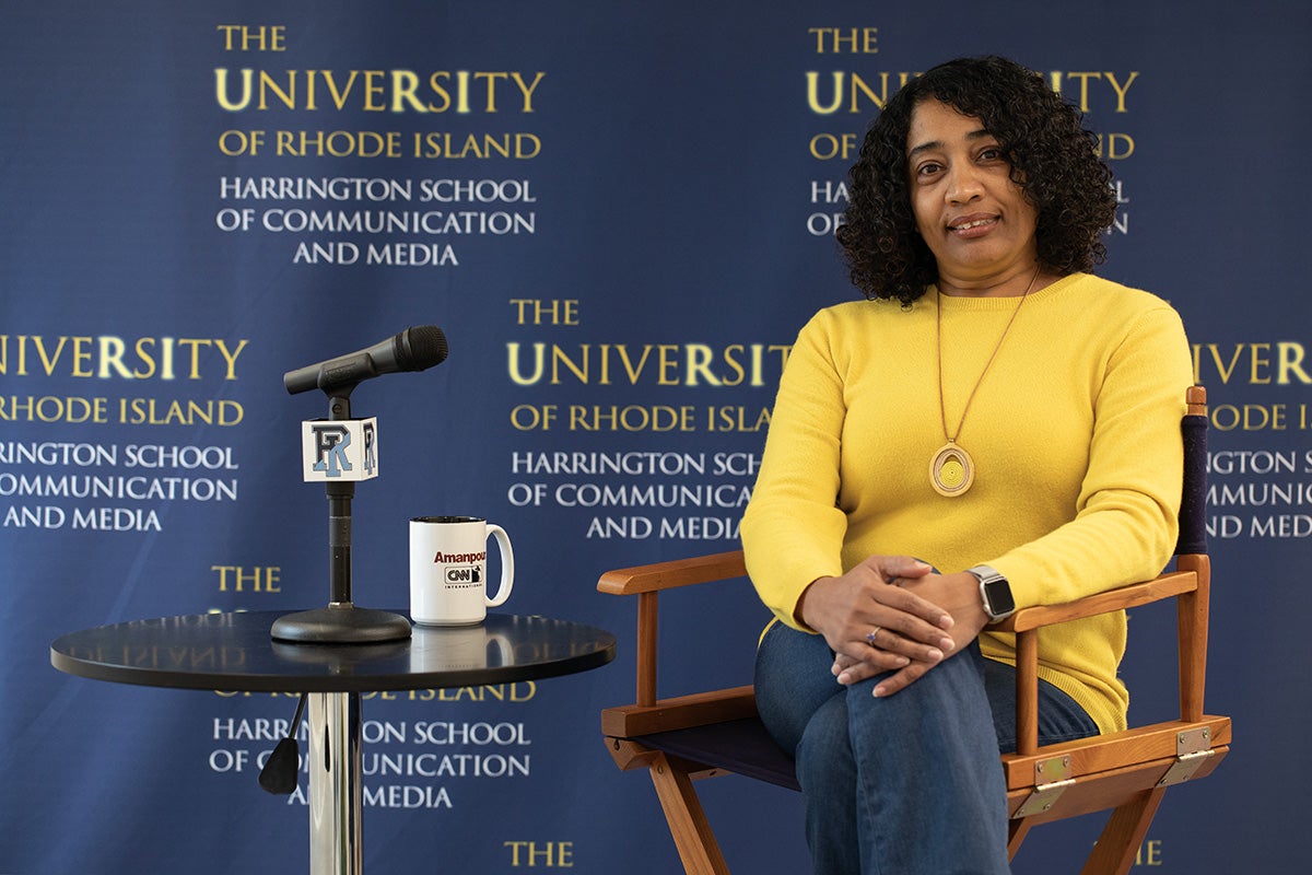 Ammina Kothari sits in a chair in profile with crossed legs wearing a yellow sweater. A table with a coffee mug is to her right.