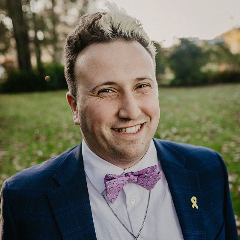 Ethan Brown wearing in profile wearing a blue blazer, a white dress shirt and purple bowtie