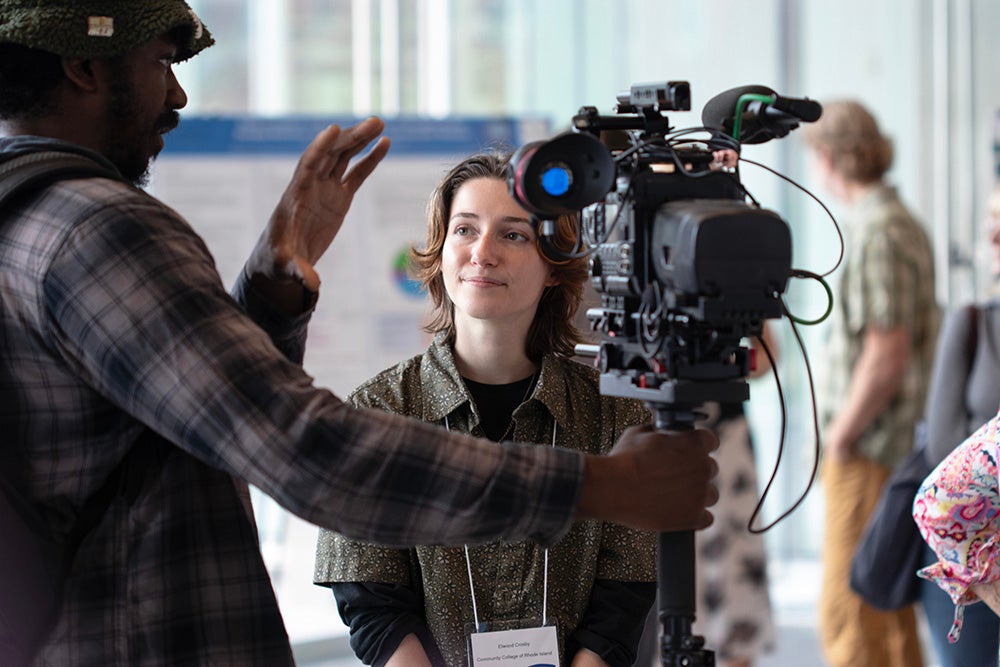 A video camera is in the foreground, held by a young man to the left wearing a flannel shirt. A young, blonde woman looks to the left listening to the young man.
