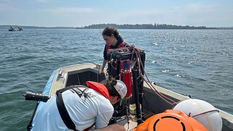 two students on a boat on the water, clear sunny day, prepare to launch a floating research device.