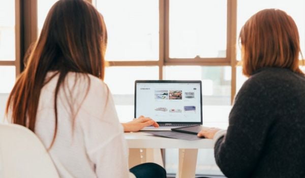 two women facing a computer and large windows