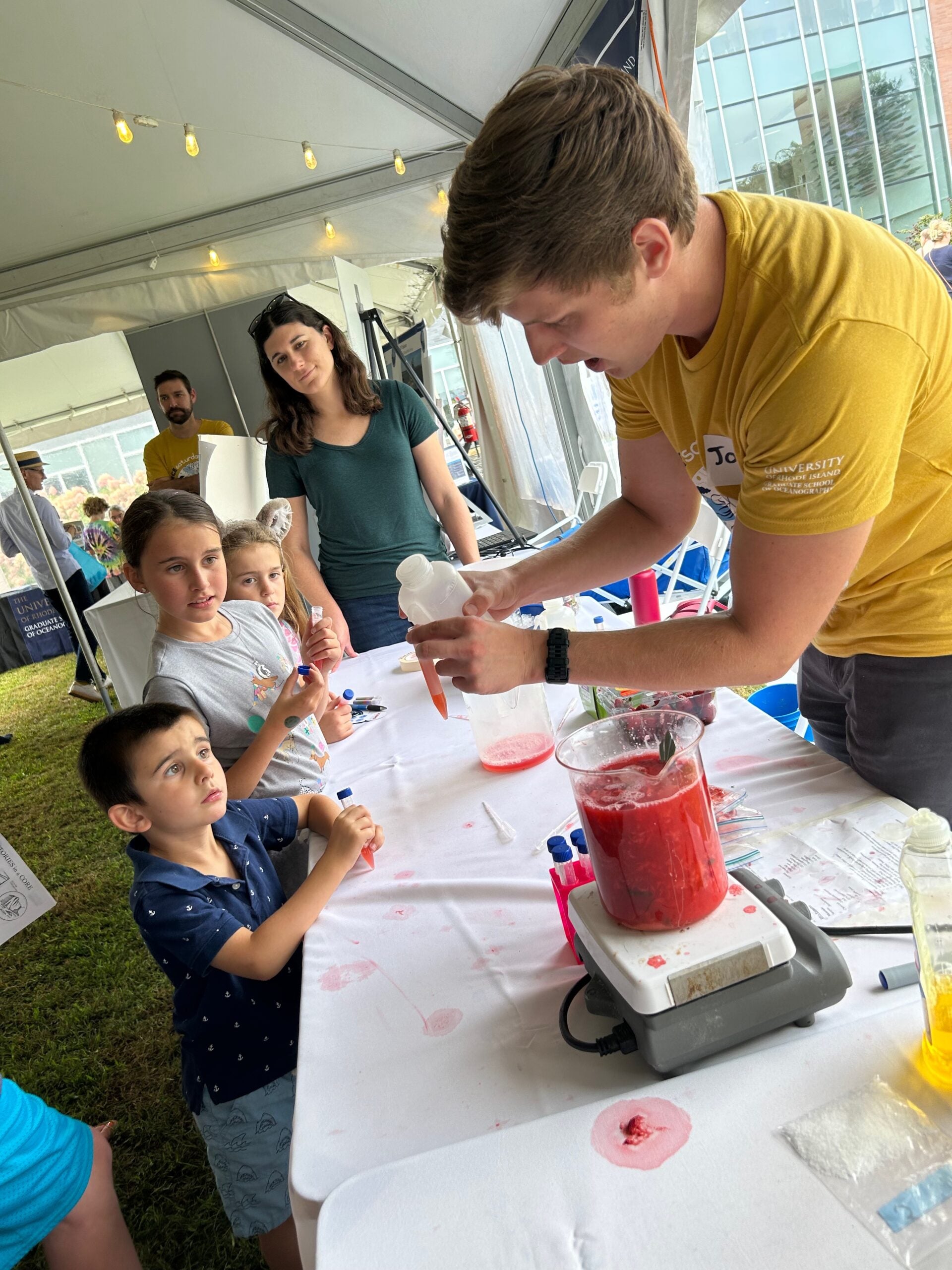 A lab member is holding a test tube to which he is adding a reagent from a squirt bottle while children watching.