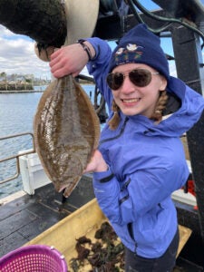 A young woman stands on a fishing boat holding up a halibut.