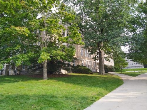 An image of Pastore Hall, a tan brick academic building with many windows. A tree and lawn are in the foreground.