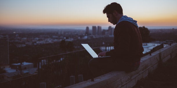 A young man works at a laptop on a balcony overlooking a city landscape.