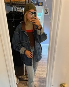 Madeline in her favorite fall sweater, a jean jacket, jeans and a green beanie taking a photo in the mirror