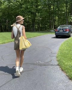 girl in left corner screen walking away towards her car in top right corner. She is carying a yellow bag, with a brown bucket hat on and green overalls with a white t-shirt underneath