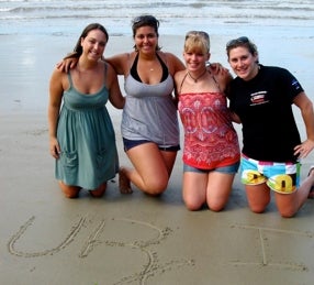 four students on the beach with URI written in the sand
