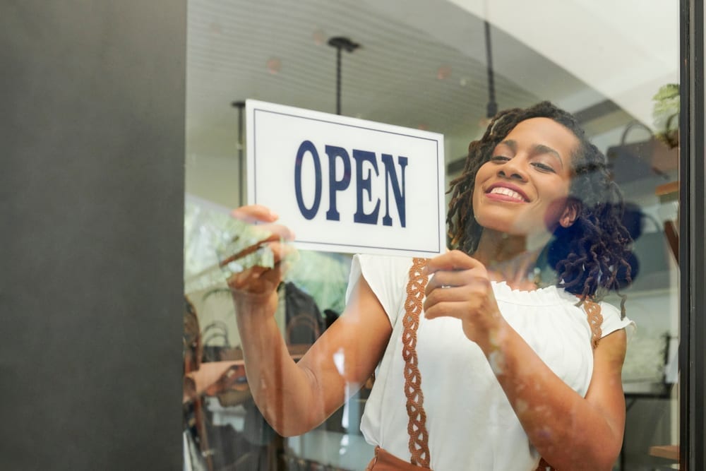 black woman in white shirt hanging an open sign in a storefront