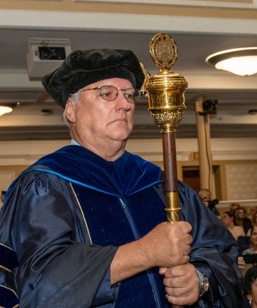 A man dressed in academic regalia carries the university mace in an inaugural procession