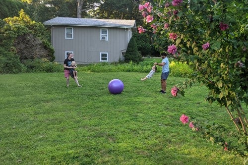 Family playing with large purple ball in yard, house in background