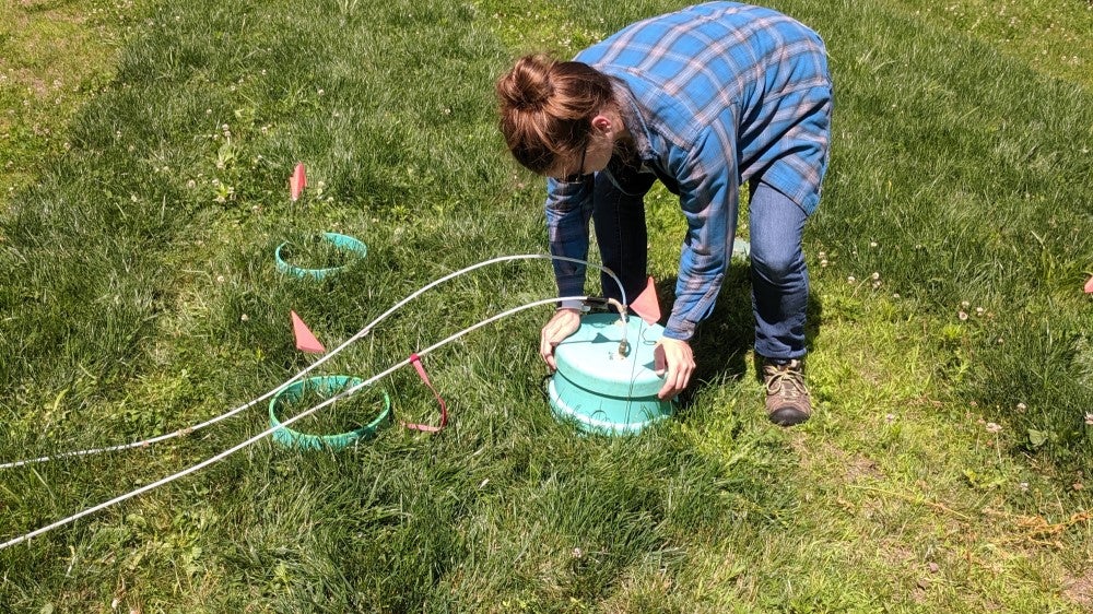 Woman bending over teal greenhouse gas sampling chamber connected to clear tubing