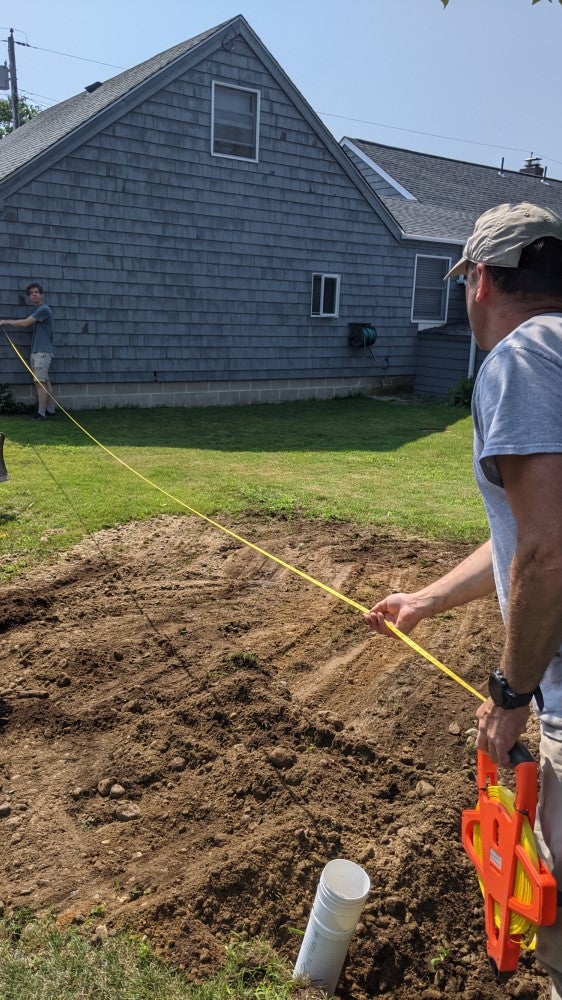 Two men measuring distance from house to white pipe in disturbed soil with yellow measuring tape