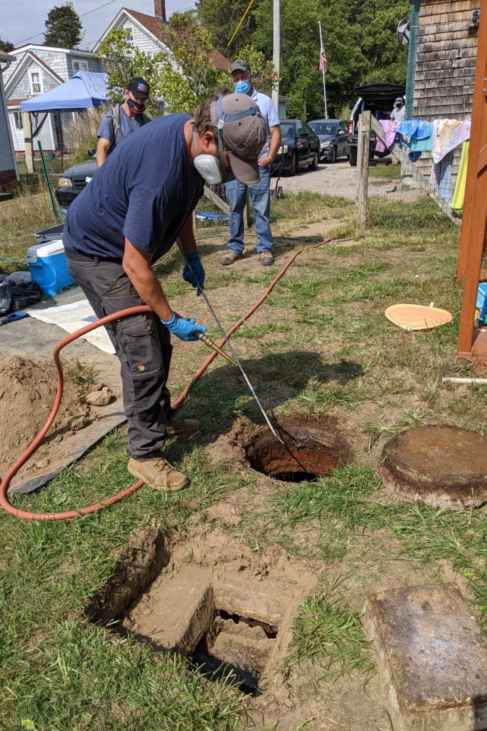 Person wearing gloves and respirator hosing tool above septic tank hole