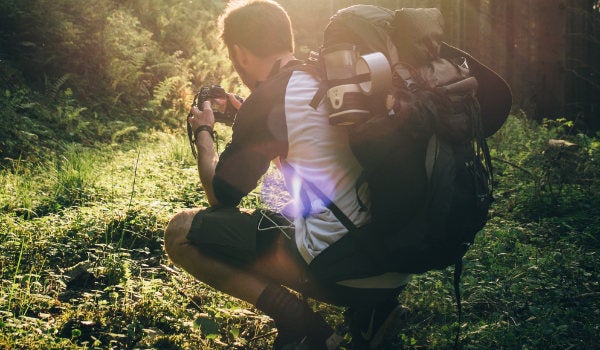 A person wearing a large backpack, in a forest setting squats to take a photograph at ground level