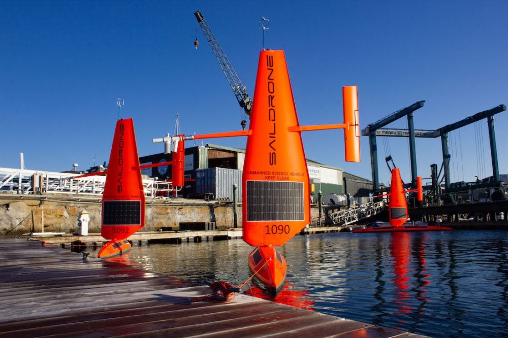 Three bright orange Saildrones (which look like robotic sailboats) are tied at a dock, a shipyard is in the background.