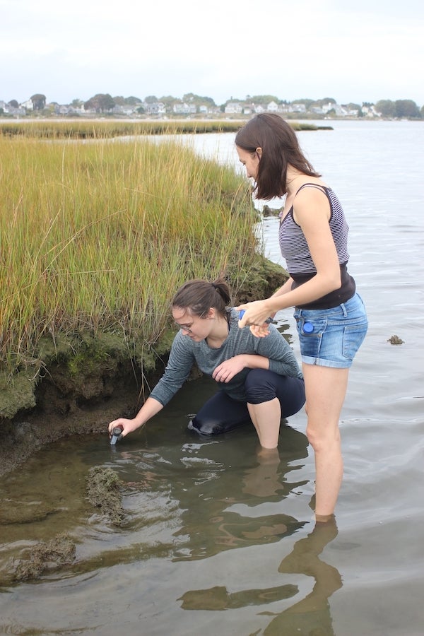 Two female graduate students outside at a salt march. One is crouched down and collecting a sample from the mud, another is standing next to her. 