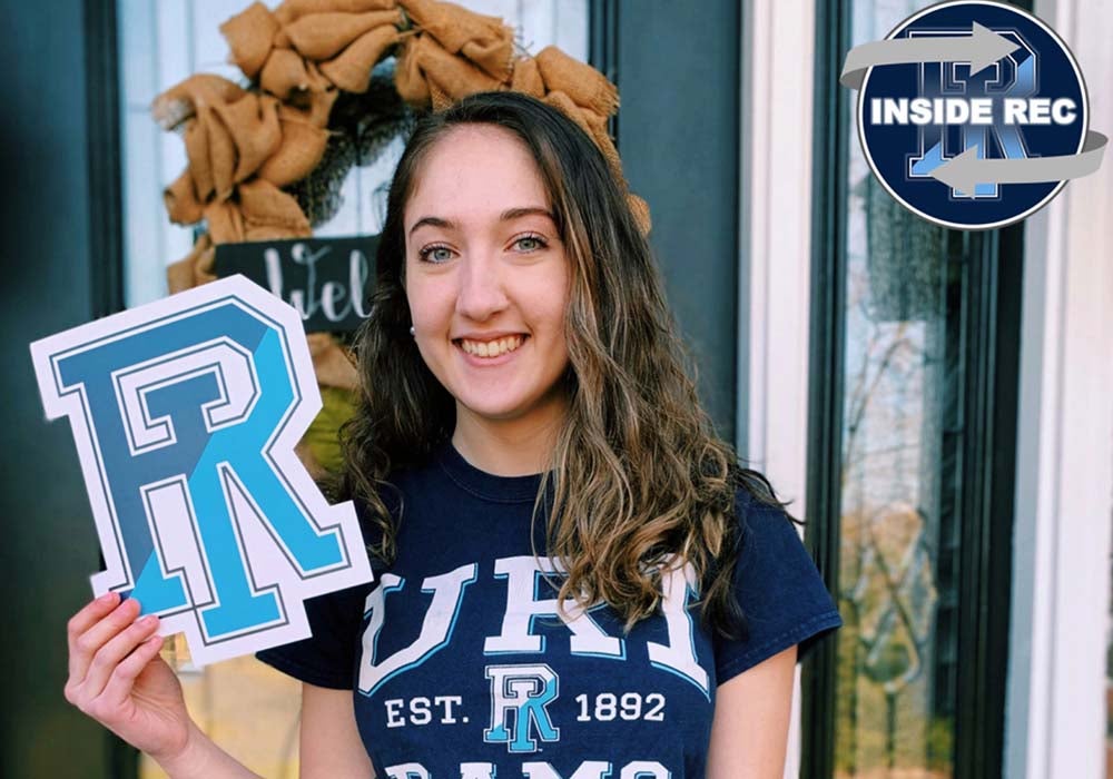 blog writer Julianna is holding a RI image in front of a door with a wreath, she is wearing a URI t-shirt