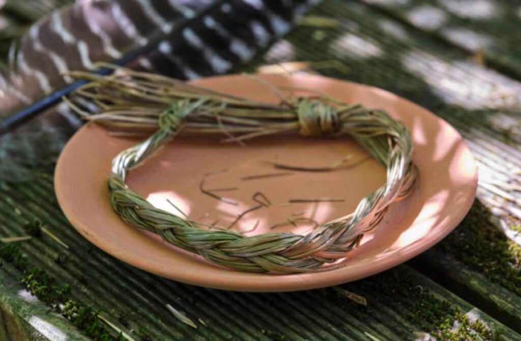 A brown ceramic plate sits in the center of the image, holding a braid of dried sweetgrass. The braided sweetgrass sits in a circular shape around the plate.